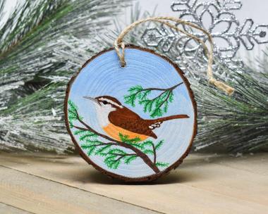 An acrylic painting of a Carolina wren perched on an evergreen tree limb is featured on a wood slice ornament at covecreekphotography.com.