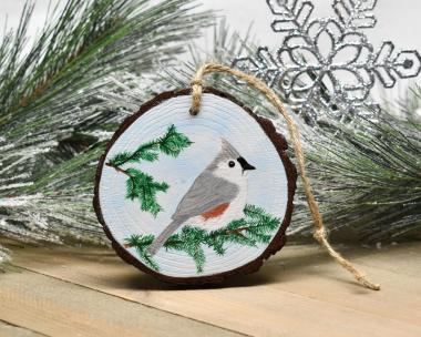 A tufted titmouse perched on an evergreen branch is hand-painted on a wood slice ornament at Cove Creek Photography.