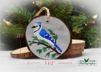 An image of a hand-painted blue jay Christmas ornament with dimensions for the wood slice at covecreekphotography.com.