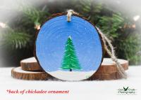 An evergreen tree in the snow on a winter day that is hand-painted on the back of a chickadee Christmas ornament at covecreekphotography.com.