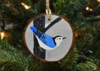 A handmade wood slice ornament with a hand-painted white-breasted nuthatch in the snow at Cove Creek Photography.