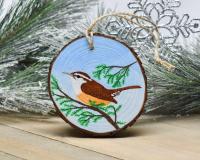 An acrylic painting of a Carolina wren perched on an evergreen tree limb is featured on a wood slice ornament at covecreekphotography.com.
