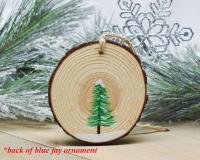 A hand-painted evergreen tree in the snow on the back of a blue jay wood slice ornament at covecreekphotography.com.