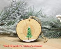 A simple evergreen tree in snow is hand-painted on the back of a cardinal wood slice Christmas ornament at covecreekphotography.com.