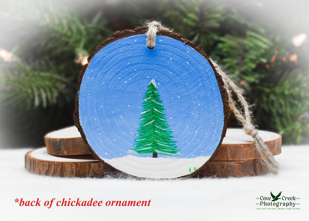 An evergreen tree in the snow on a winter day that is hand-painted on the back of a chickadee Christmas ornament at covecreekphotography.com.