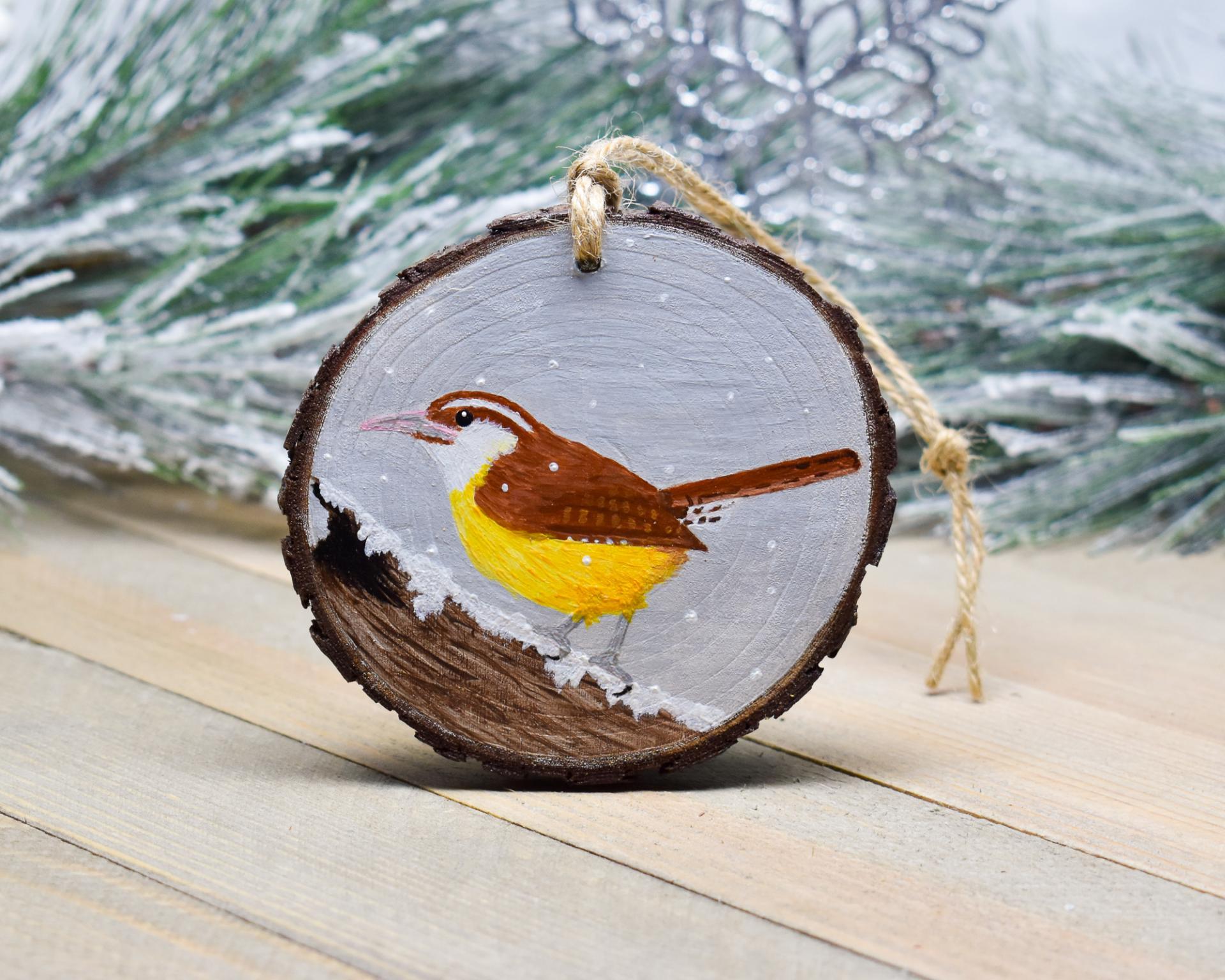 A handmade woos slice Christmas ornament with a hand-painted Carolina wren on a snow covered log at covecreekphotography.com.