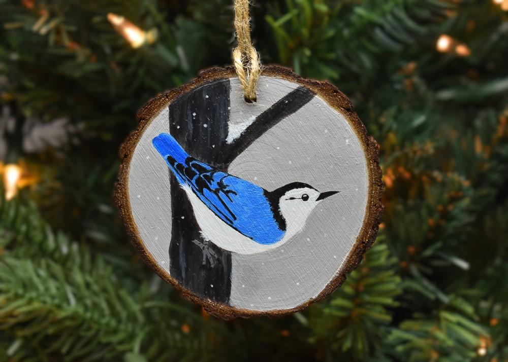 A handmade wood slice ornament with a hand-painted white-breasted nuthatch in the snow at Cove Creek Photography.