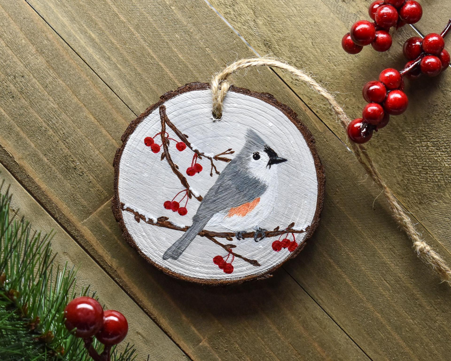 An acrylic painting of a tufted titmouse perched among tree branches and red berries on a wood slice ornament at covecreekphotography.com.