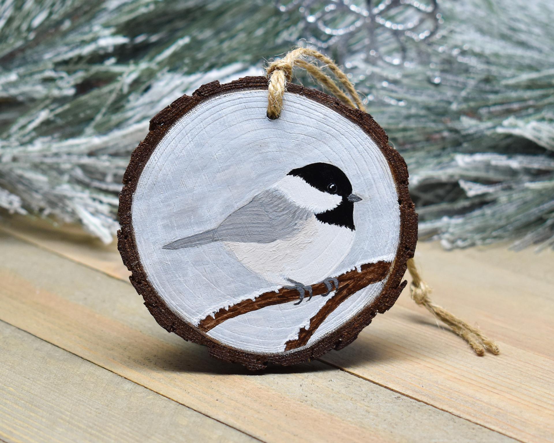 A wood slice Christmas ornament with a hand-painted black-capped chickadee perched on a snow covered branch at covecreekphotography.com.