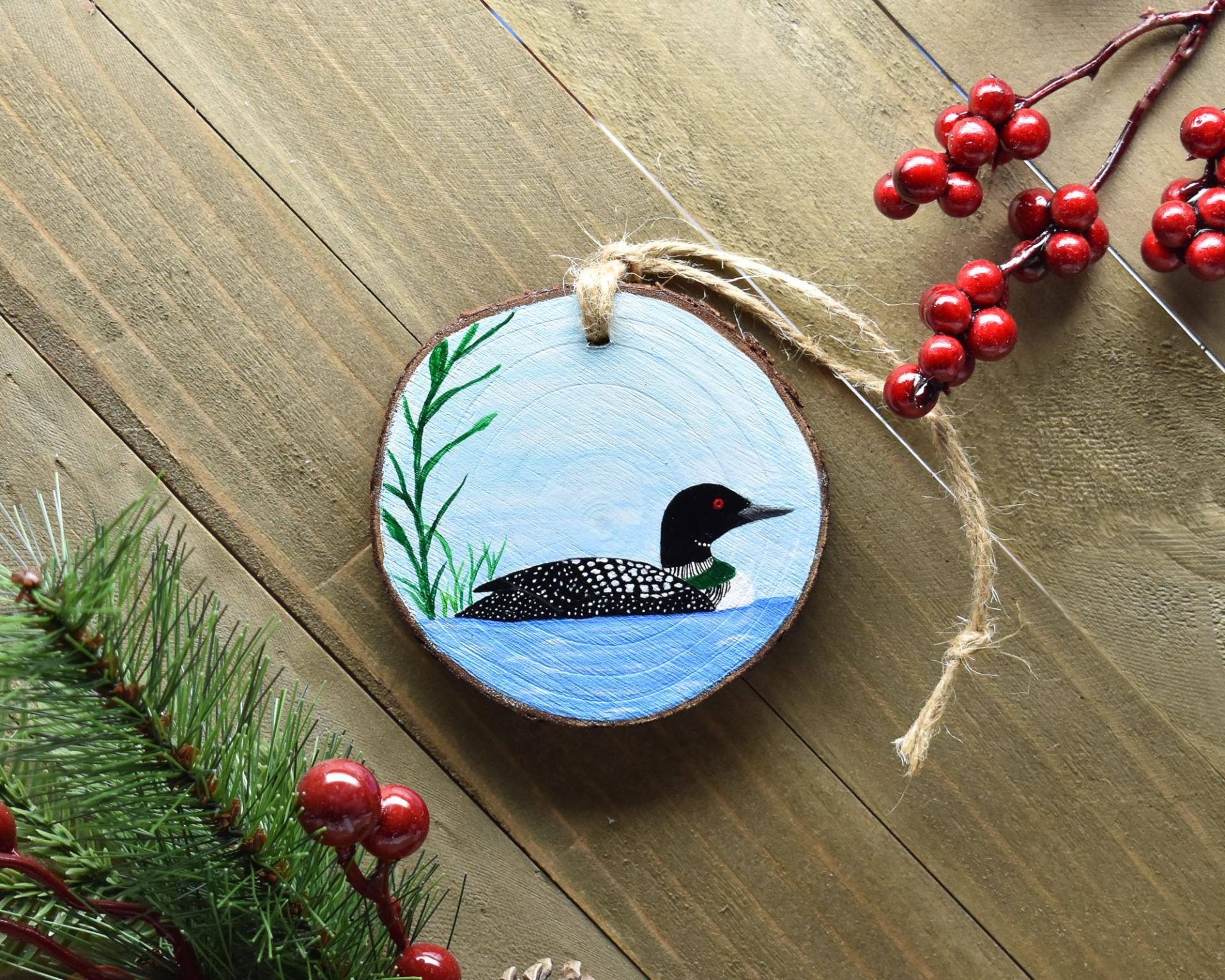 A hand-painted common loon on a pond wood slice Christmas ornament is displayed on a wooden board at Cove Creek Photography.
