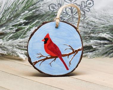 A red cardinal on a snow dusted branch with a sky background that is hand-painted on a wood round at Cove Creek Photography.