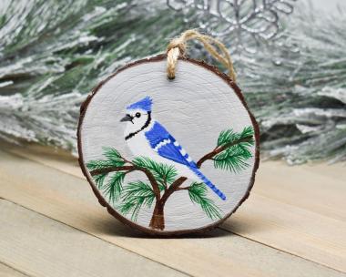 A hand-painted wood slice Christmas ornament with a blue jay perched in the top of a cedar tree with a winter gray background at Cove Creek Photography.