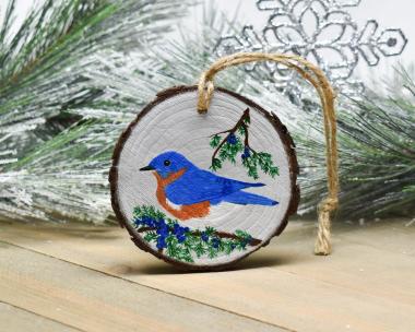 A hand-painted blue jay amid cedar branches and blue berries on a wood slice Christmas ornament at Cove Creek Photography.