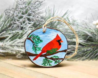 A birght red male northern cardinal with blue sky background is hand-painted on a wood slice ornament at covecreekphotography.com.